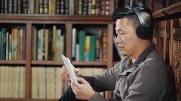 Homme asiatique dans les écouteurs bénéficie d'une tablette dans la bibliothèque. Sur fond d'étagères avec livres — Video
