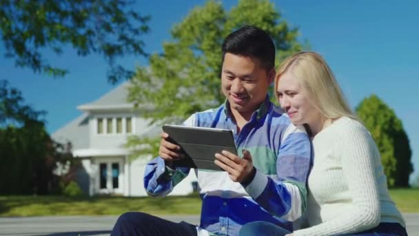 Feliz jovem casal desfrutando de um tablet. Sentam-se ao lado da casa. — Vídeo de Stock