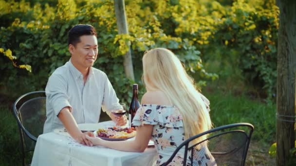 Счастливый азиат ужинает с красивой женщиной в элегантном летнем платье. Сидеть за столом в винограднике, пить вино — стоковое видео