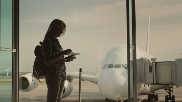 Una silueta de una mujer con documentos de embarque en la mano, esperando aterrizar en su vuelo. Se para en la ventana de la terminal del aeropuerto —  Fotos de Stock