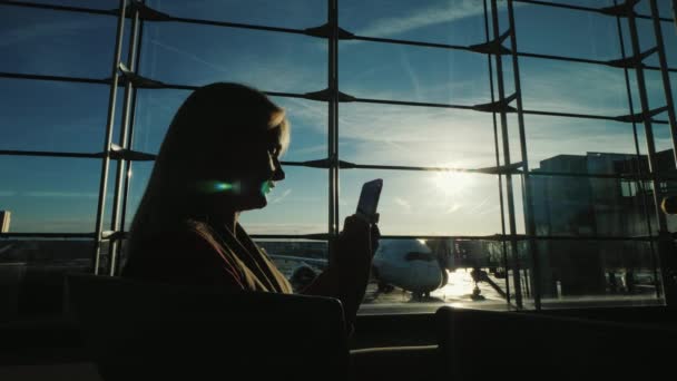 Silueta mladé ženy s chytrým telefonem. Sedí na velké okno na letištní terminál — Stock video