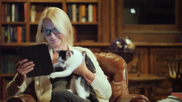 Eine Frau liest die Nachrichten auf einem Tablet, sitzt zu Hause in einer soliden Bibliothek, in ihrem Arm ein verspieltes Kätzchen. Komfort mit einem Haustier — Stockvideo