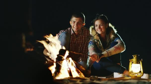 Het gezelschap van vrienden ontspannen rond het kampvuur, geroosterde marshmallows op stokken boven een vuur — Stockvideo