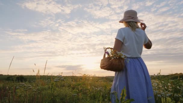 夕日を楽しみにして、草原の中と野生の花のバスケットの帽子の中年の女性が立っています。 — ストック動画