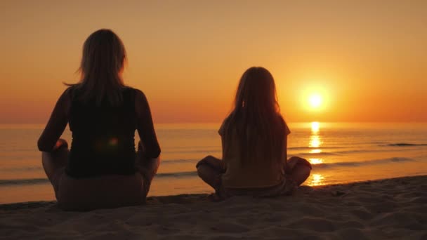 Egy nő a lányával egymás mellett ült a homokon, Lótusz póz, nézi a naplementét a tenger felett — Stock videók