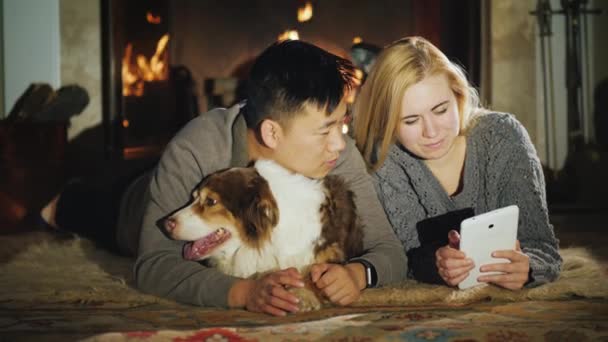 Una pareja multiétnica se encuentra cerca de la chimenea, utilizando una tableta. Junto a ellos está su perro - Australian Shepherd — Vídeo de stock