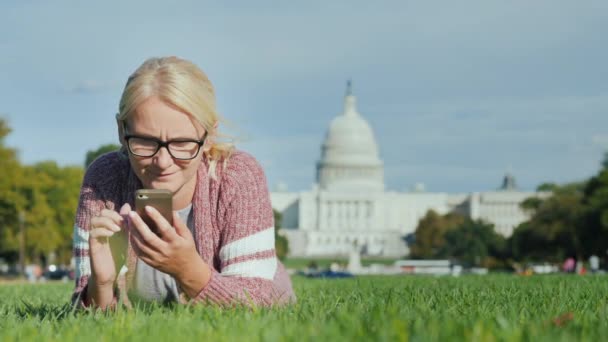 Mujer joven descansando en un césped verde en el fondo del Capitolio en Washington. Utiliza un teléfono inteligente — Vídeo de stock