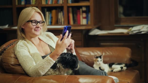 La donna usa lo smartphoneat a casa, ha un cucciolo tra le braccia e sullo sfondo un gatto gioca con la coda. Accoglienza e armonia con gli animali domestici — Video Stock