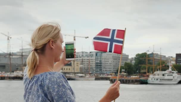Visão traseira: Uma jovem com uma bandeira norueguesa fotografa-se no fundo da linha da cidade de Oslo. Viagem Escandinávia e Noruega conceito — Vídeo de Stock