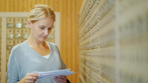 Una mujer de mediana edad con el pelo rubio está de pie en la oficina de correos al lado de las celdas individuales y mirando las cartas recibidas — Vídeos de Stock