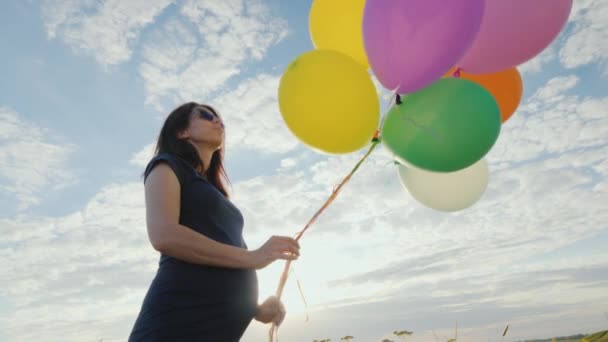 Carefree pregnant woman playing with balloons in a meadow, against a blue sky background — Stock Video