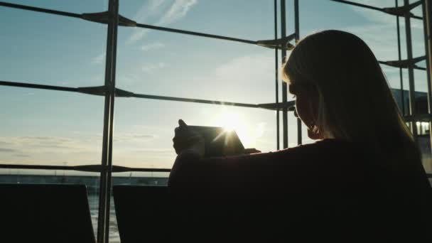 Conectado en un viaje. Una mujer mirando la pantalla de una tableta, se sienta frente a una gran ventana en la terminal del aeropuerto — Vídeo de stock