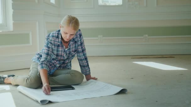 Γυναίκες σχεδιαστής εργάζεται με ένα σχέδιο, κάθεται στο πάτωμα σε ένα ευρύχωρο δωμάτιο ενός σπιτιού νέα, ημιτελής. — Αρχείο Βίντεο