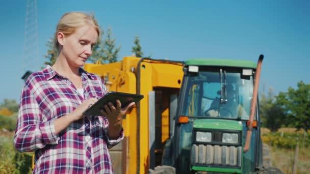 Een vrouwelijke boer maakt gebruik van een tablet op het veld, op de achtergrond is een trekker — Stockvideo