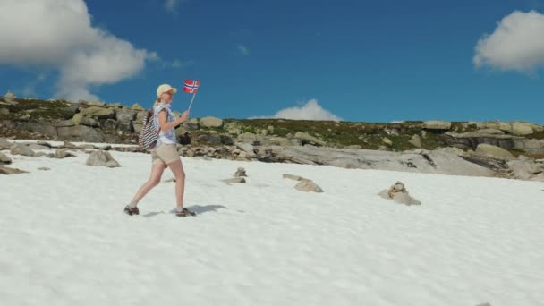 Una donna cammina nella neve che non si è sciolta in montagna in estate. Tiene in mano la bandiera della Norvegia — Video Stock