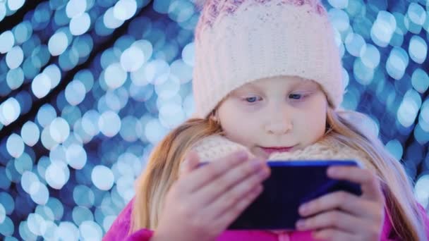 Un niño con ropa de invierno rosa usa un teléfono inteligente afuera contra las luces borrosas de una guirnalda festiva — Vídeos de Stock