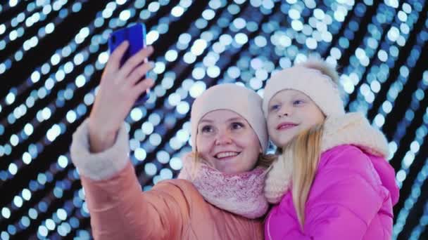 Мама и дочь фотографируются на фоне размытых праздничных гирлянд в городе — стоковое видео