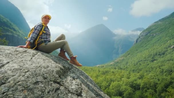 若い女性が背の高い山に囲まれた大きな岩の上に座っている。信じられないほど美しい自然の中で — ストック動画
