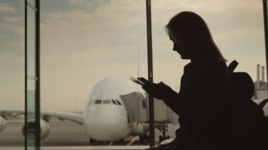 Terminalin penceresinde elinde biniş belgeleriyle duran bir kadının silüeti. Pencerenin dışında güzel bir uçak var.