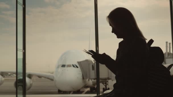 Силует жінки з посадковими документами, що стоять у вікні терміналу. Над вікном красивий авіалайнер — стокове відео