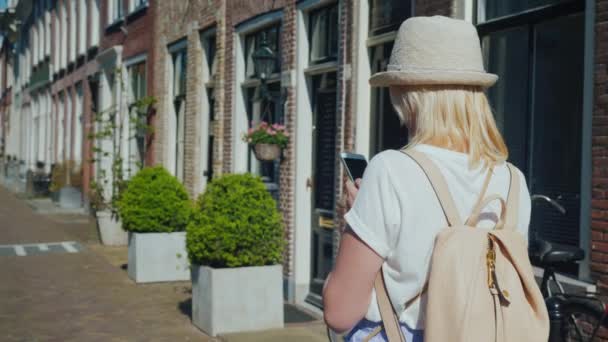 一个戴着帽子和背包的女人沿着荷兰代尔夫特古城的狭窄街道走去 使用智能手机 欧洲旅游概念 斯坦尼康射击 — 图库视频影像