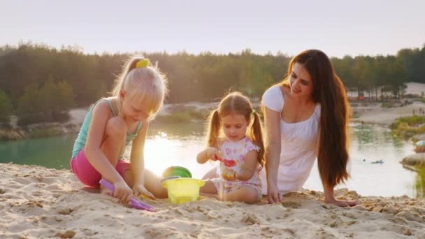 Joven madre juega en la arena con dos hijas. Al atardecer en el fondo del hermoso lago — Vídeo de stock