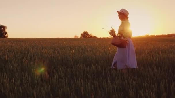 Молода жінка з кошиком квітів, що гуляє полем на заході сонця — стокове відео