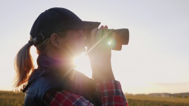 Ung kvinna i en mössa ser genom kikare. Solen skiner genom hennes händer — Stockvideo