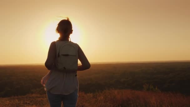 Visão traseira: Uma jovem mulher com uma mochila corre para a frente em direção ao sol. Cabelo brilha nos raios do pôr do sol — Vídeo de Stock