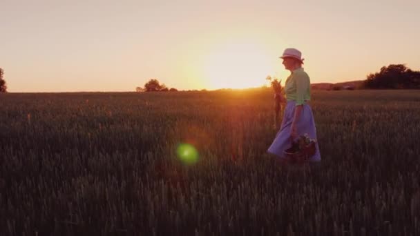 サイドビュー: 野生の花のバスケットと幸せな屈託のない女は美しい場所で夕暮れ歩く — ストック動画