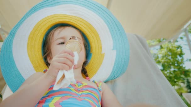 Una niña en un gran sombrero disfruta comiendo helado con placer . — Vídeo de stock