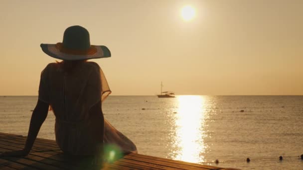 Een vrouw zit op een houten pier, voldoet aan de dageraad van de zee. Een schip is zichtbaar in de verte. Dromen en reizen concept — Stockvideo