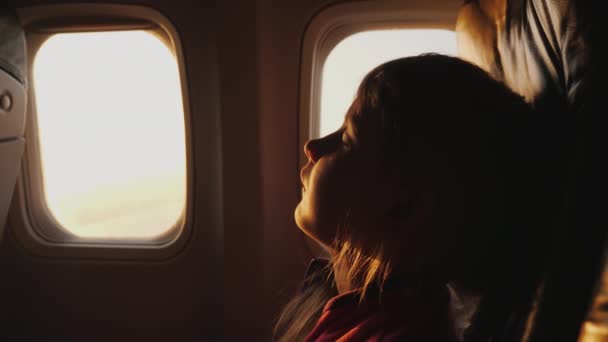 Спати будь-де і в будь-якому положенні, маленька дівчинка спить в кріслі на борту літака, ранкове світло пробивається крізь вікно літака — стокове відео