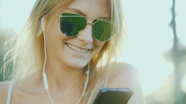 Stylish young woman listening to music on headphones, holding a smartphone in her hand. — Stock Video