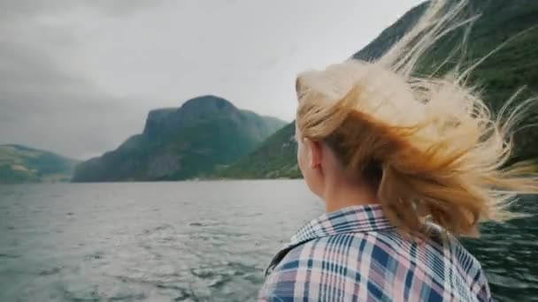 Kobieta z długimi włosami stoi na dziobie statku wycieczkowego, podziwiając fiord w Norwegii. Wiatr powiewa jej włosy — Wideo stockowe