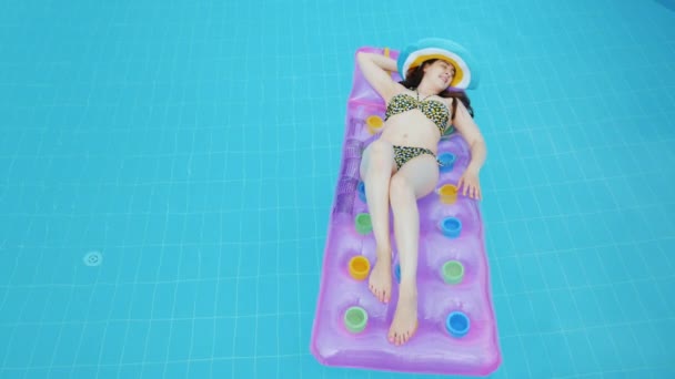 En ung flicka med mörkt hår i en färgglad snygg hatt solbad på madrassen i poolen. — Stockvideo