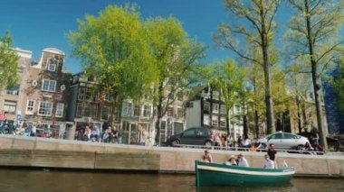 Amsterdam kanal boyunca yelken. Güzel evleri, park etmiş arabaların bir satırı, güneşin görünümü güzel pencerelerde yansıtılır. Turizm Hollanda. 4k video