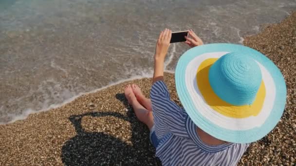 As impressões brilhantes de umas férias de praia na praia com cascalho, uma menina turística em um chapéu brilhante luxuoso com campos grandes fazem uma foto em um telefone celular — Vídeo de Stock