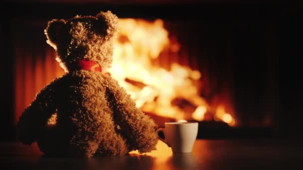 Um filhote de urso com uma xícara de chá fica em frente à lareira. Conforto e calor na casa — Vídeo de Stock