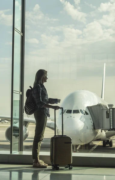 Donna con carte d'imbarco e bagaglio a mano guarda fuori dalla finestra del terminal su un grande aereo di linea — Foto Stock