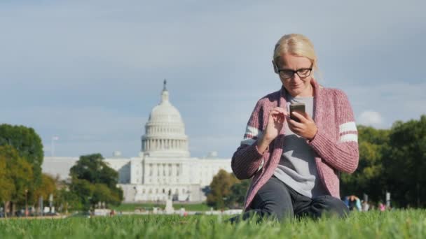 Młoda kobieta w okularach siedzi na trawniku, cieszy się smartfonem. Na tle Kapitolu w Waszyngtonie, DC — Wideo stockowe