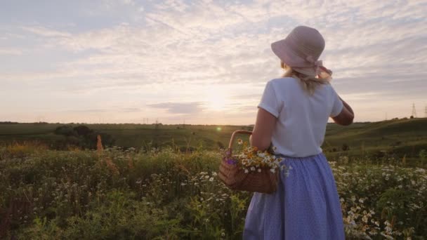 Una mujer de mediana edad en un sombrero con una cesta de flores silvestres se encuentra en un prado, mirando hacia el atardecer — Vídeos de Stock