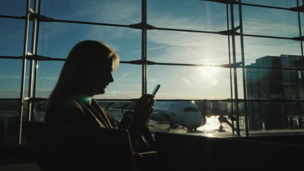 Silhouet van een vrouw, die zit bij het raam in de terminal van de luchthaven, met behulp van een smartphone — Stockvideo