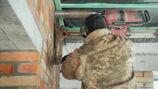 En man i skyddande hörlurar borrar en tegel vägg för att lägga ventilations rör. — Stockvideo