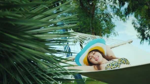 Una joven feliz en un sombrero de ala ancha brillante y en un bikini se balancea en una hamaca cerca de un bassen abierto con palmas verdes — Vídeos de Stock