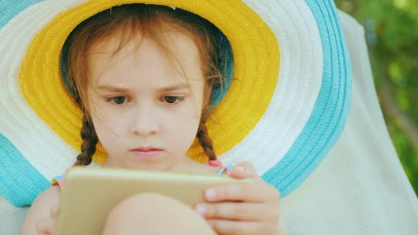 Roztomilá holčička v velkým pestrobarevným klobouku v plavkách sedí na lehátku a hraje na její mobilní telefon — Stock video