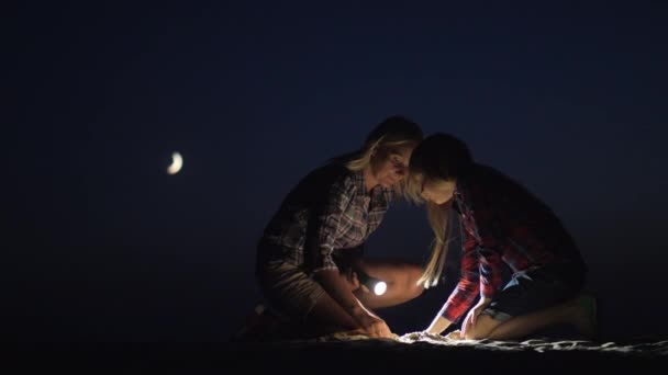 Mamma e figlia giocano insieme nella sabbia di notte. Brillano con una torcia alla ricerca di qualcosa. Avventura e un momento felice insieme — Video Stock