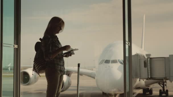 Una silueta de una mujer con documentos de embarque en la mano, esperando aterrizar en su vuelo. Se para en la ventana de la terminal del aeropuerto — Vídeo de stock