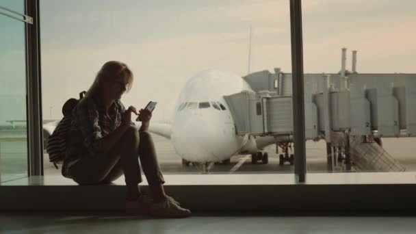 Женщина с телефоном в руке сидит на подоконнике в терминале аэропорта на фоне авиалайнера за окном — стоковое видео