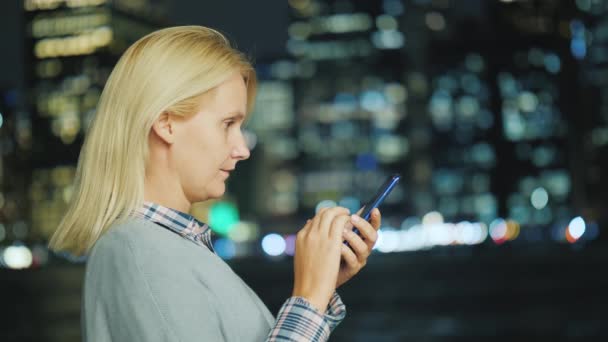 Mujer blanca de mediana edad utiliza un teléfono inteligente en el fondo de las luces de Manhattan — Vídeos de Stock